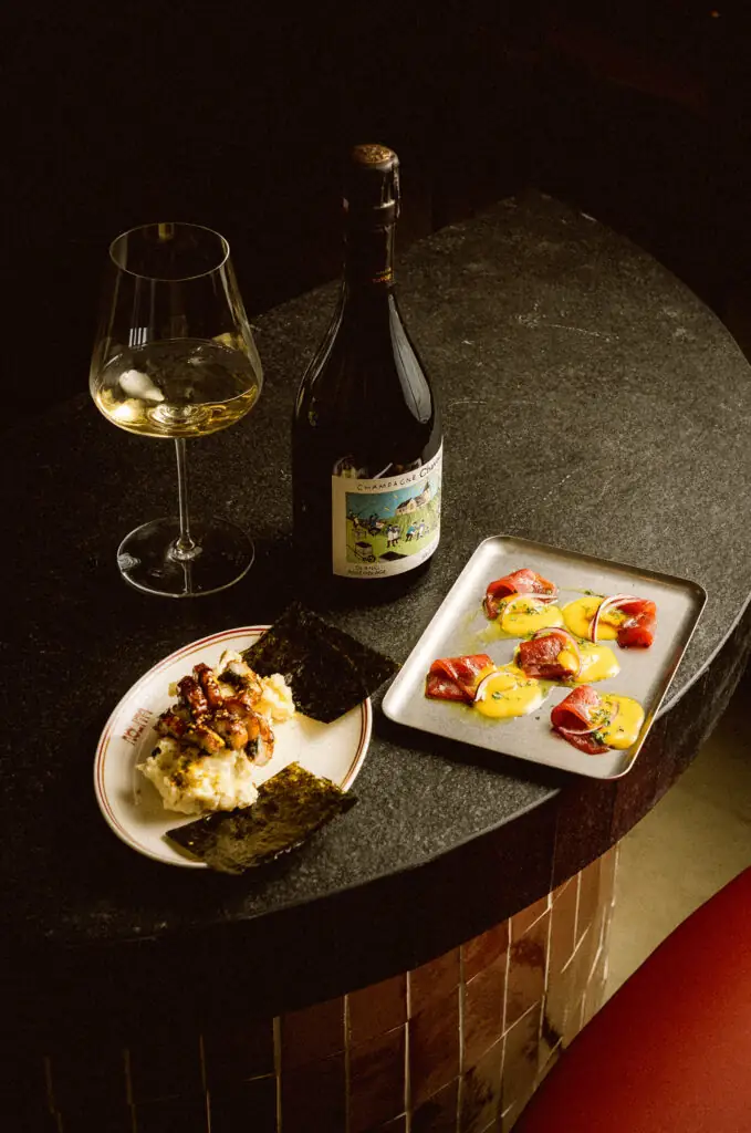 Dark round table with a glass of white wine, a champagne bottle, a plate of grilled skewers on mash with nori, and a tray of tuna in yellow sauce