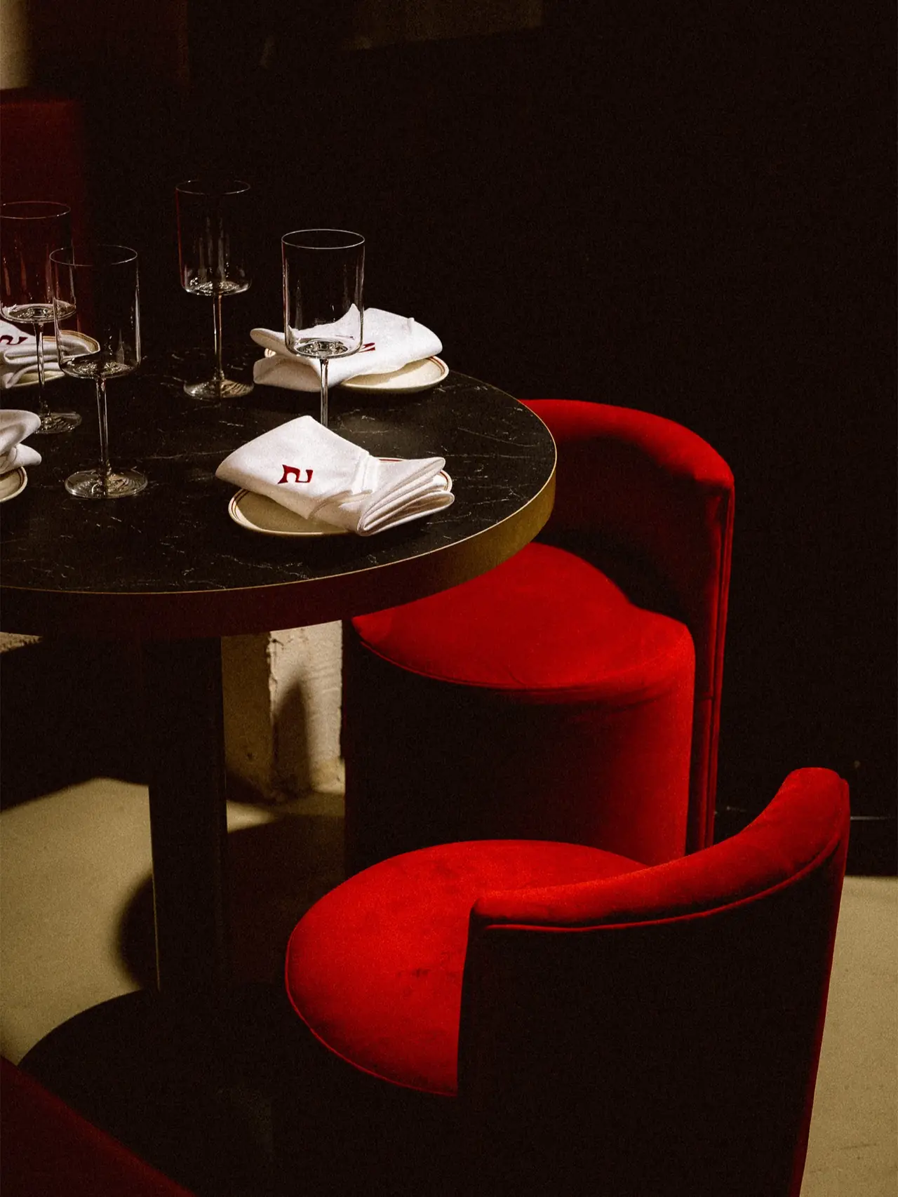 Set black marble bistro table with wine glasses and monogrammed white napkins on plates, flanked by red velvet tub chairs