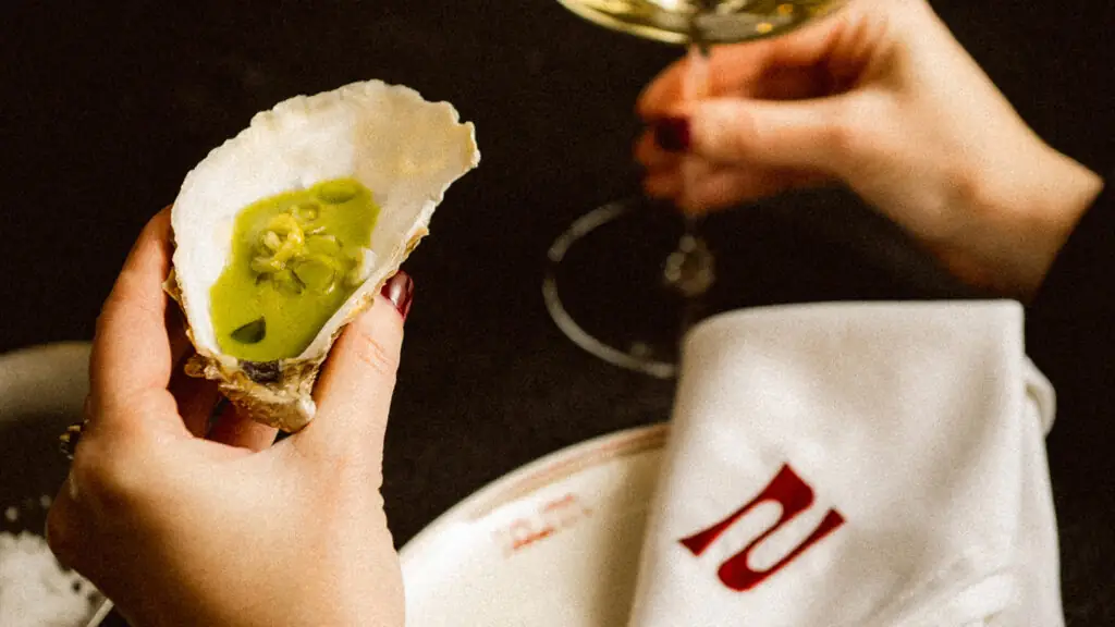 Hand holding an oyster on the half shell filled with green mignonette, with a second hand raising a wine glass and a monogrammed napkin below