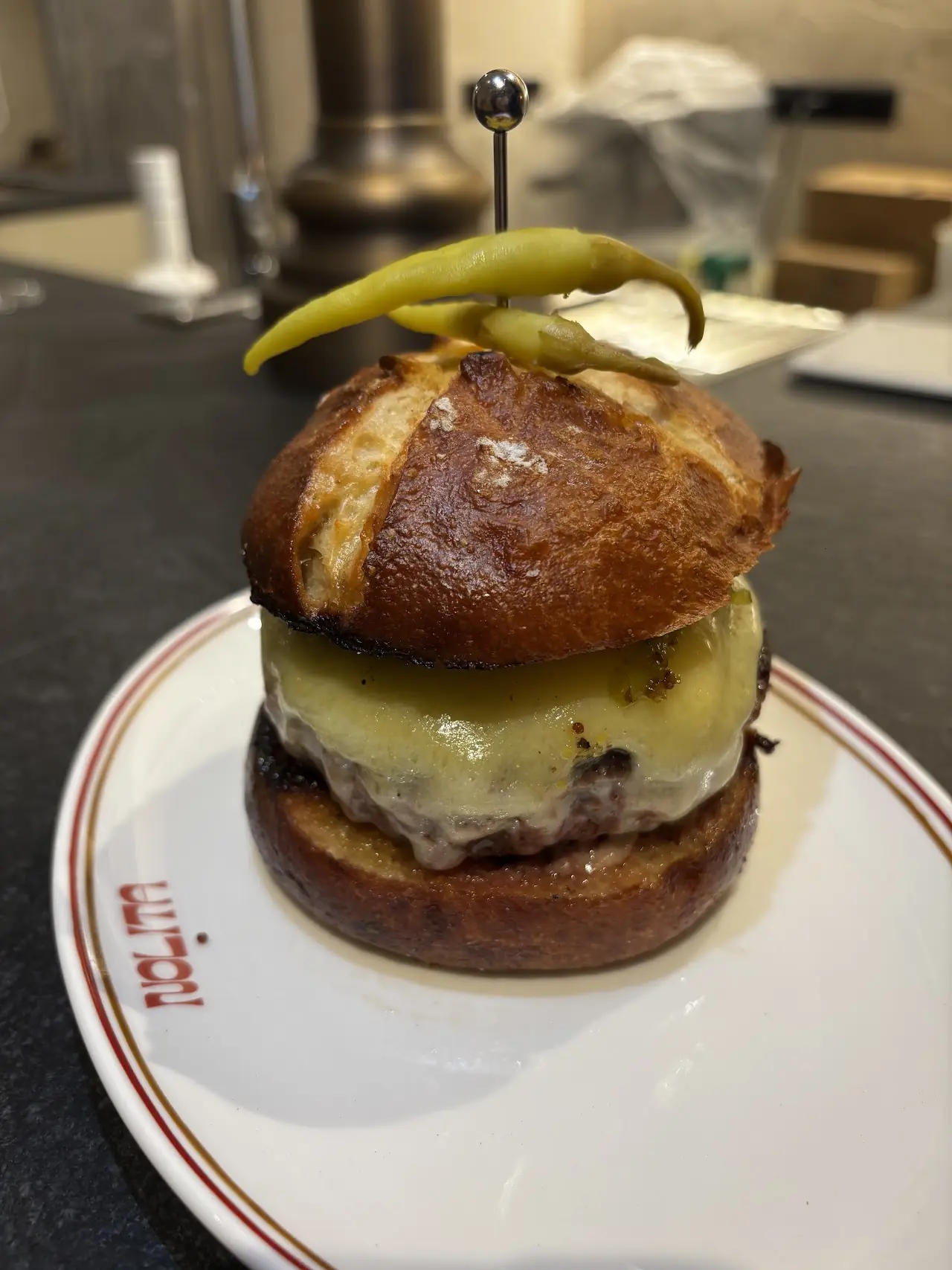 A stacked burger with melted cheese on a pretzel-style bun, topped with a pickled chili, on a white plate.