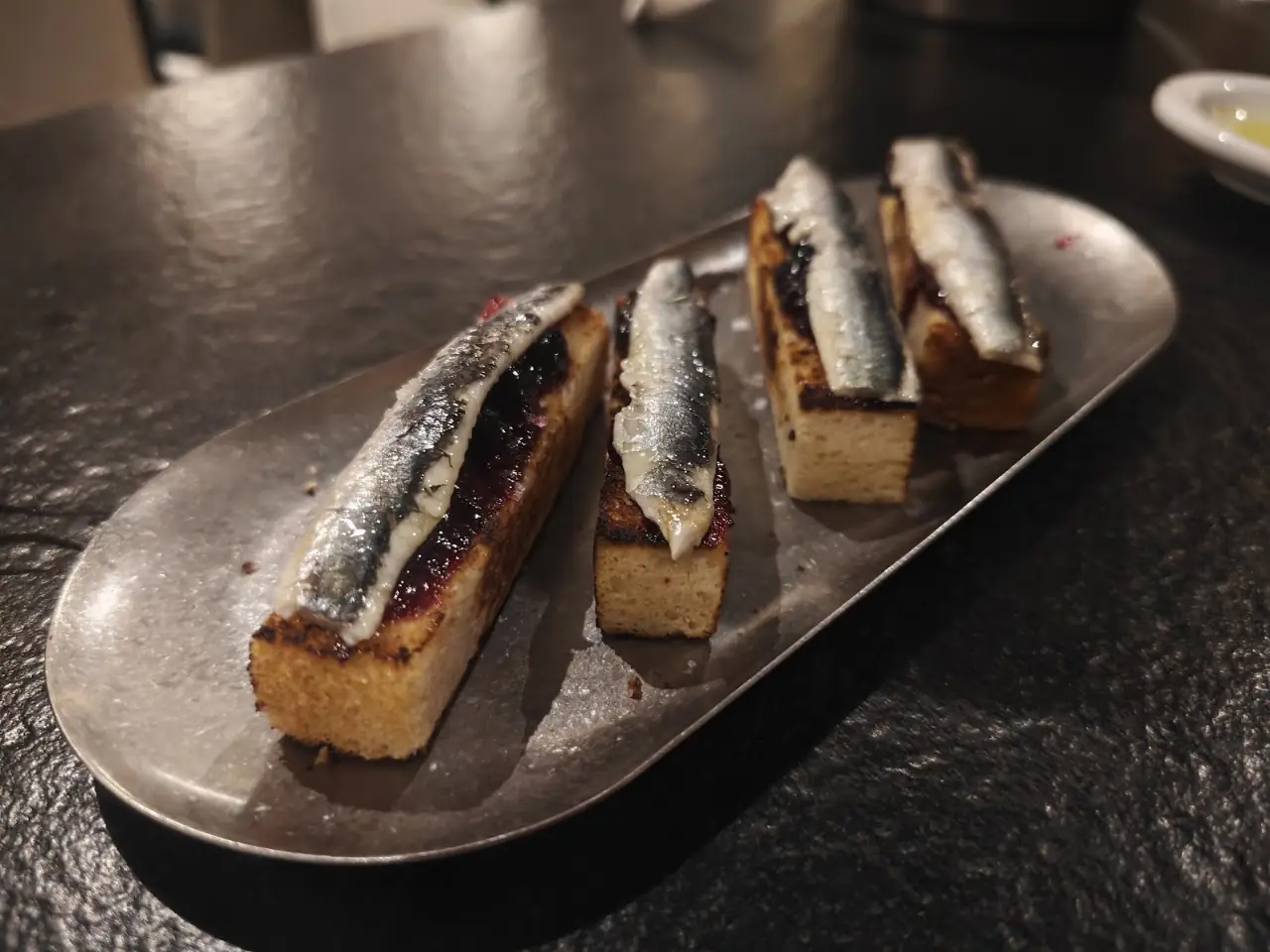 Four grilled sardine fillets on toasted bread slices, arranged on an oval metal plate.