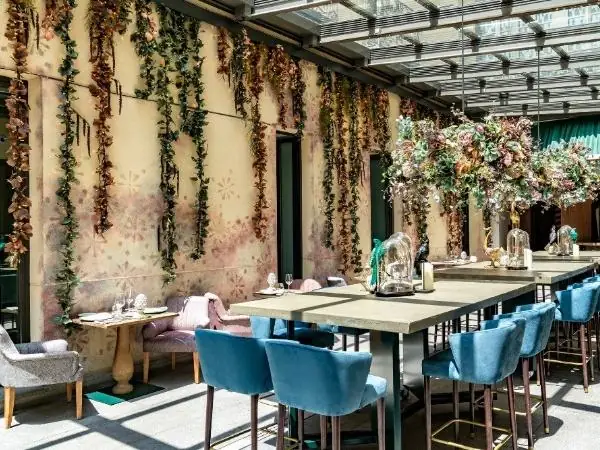 Bright glass-roofed dining space with hanging greenery, blue velvet chairs, floral centrepieces and a painted wall