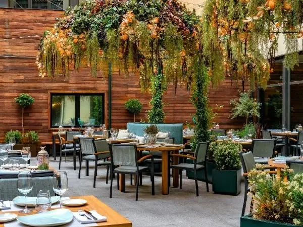 Courtyard terrace dining area with lush overhead hanging plants, wooden tables and grey chairs against a timber wall
