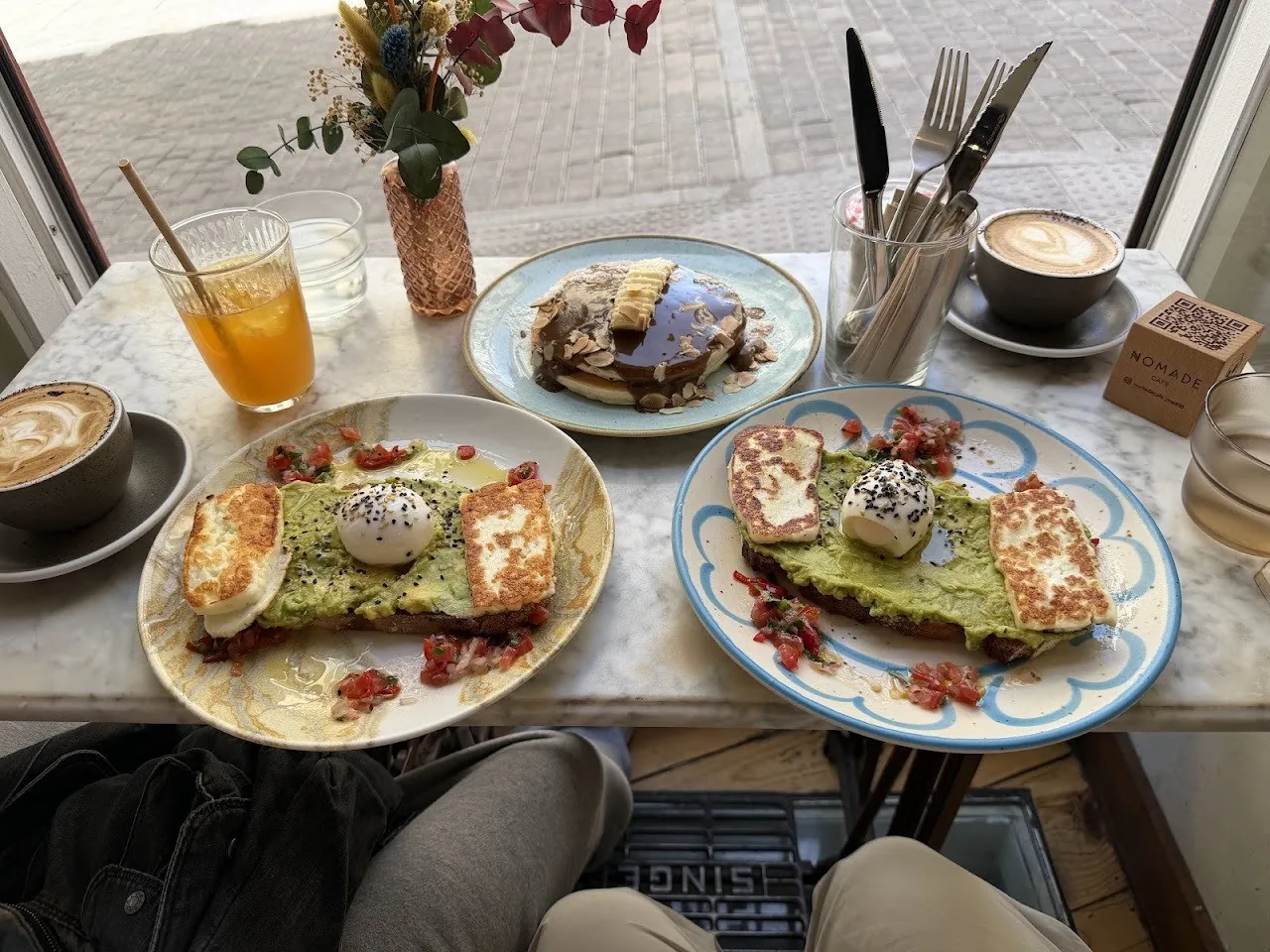 Two plates of avocado toast topped with eggs, with a coffee, orange juice and a small plate of pancakes on a marble table