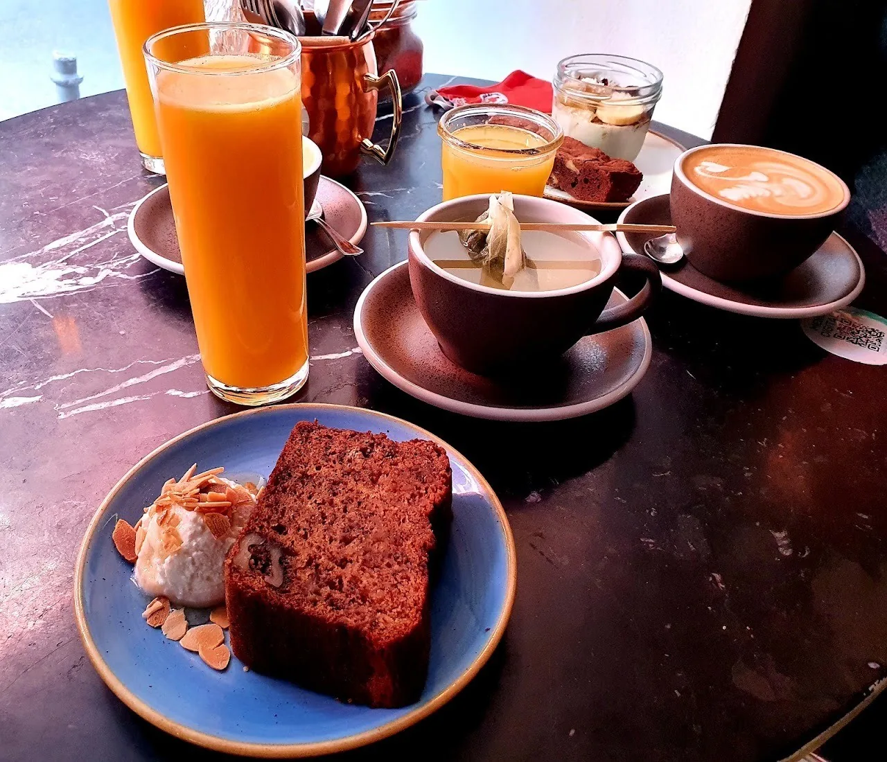 A slice of cake on a blue plate next to a glass of orange juice and two coffees