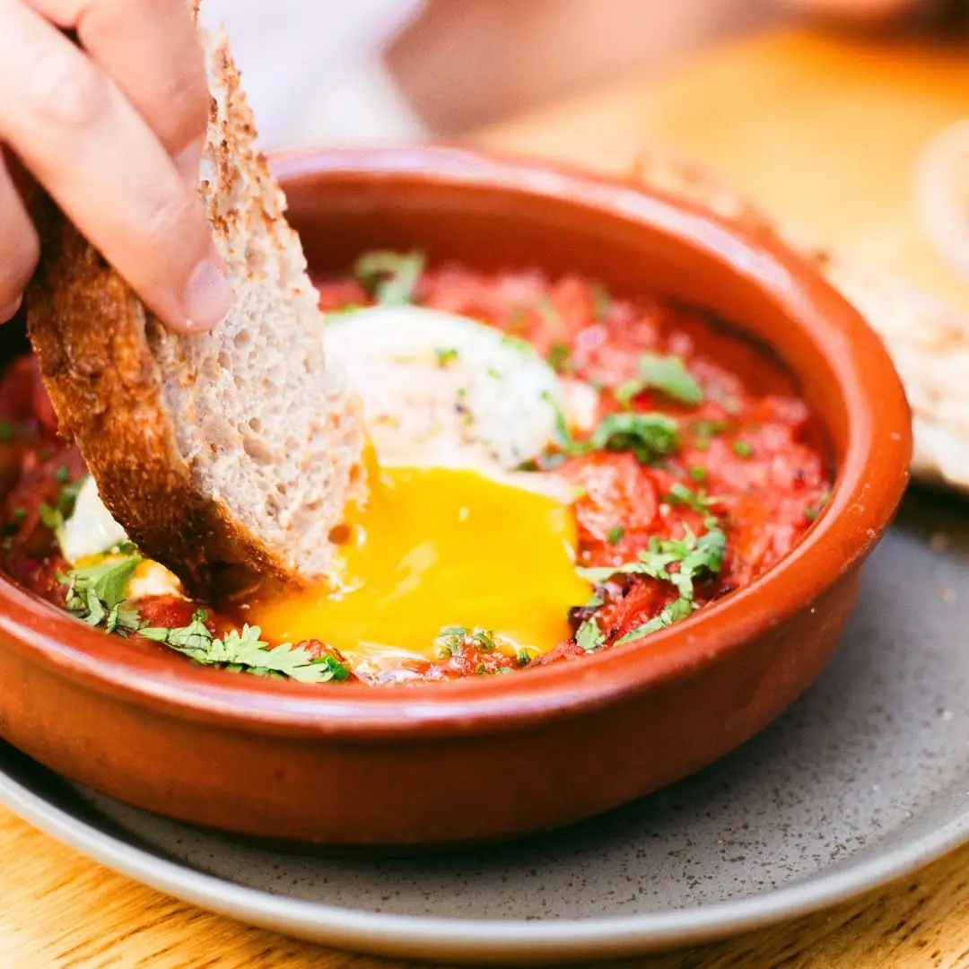 Bread being dipped into a shakshuka with a runny egg in a terracotta dish