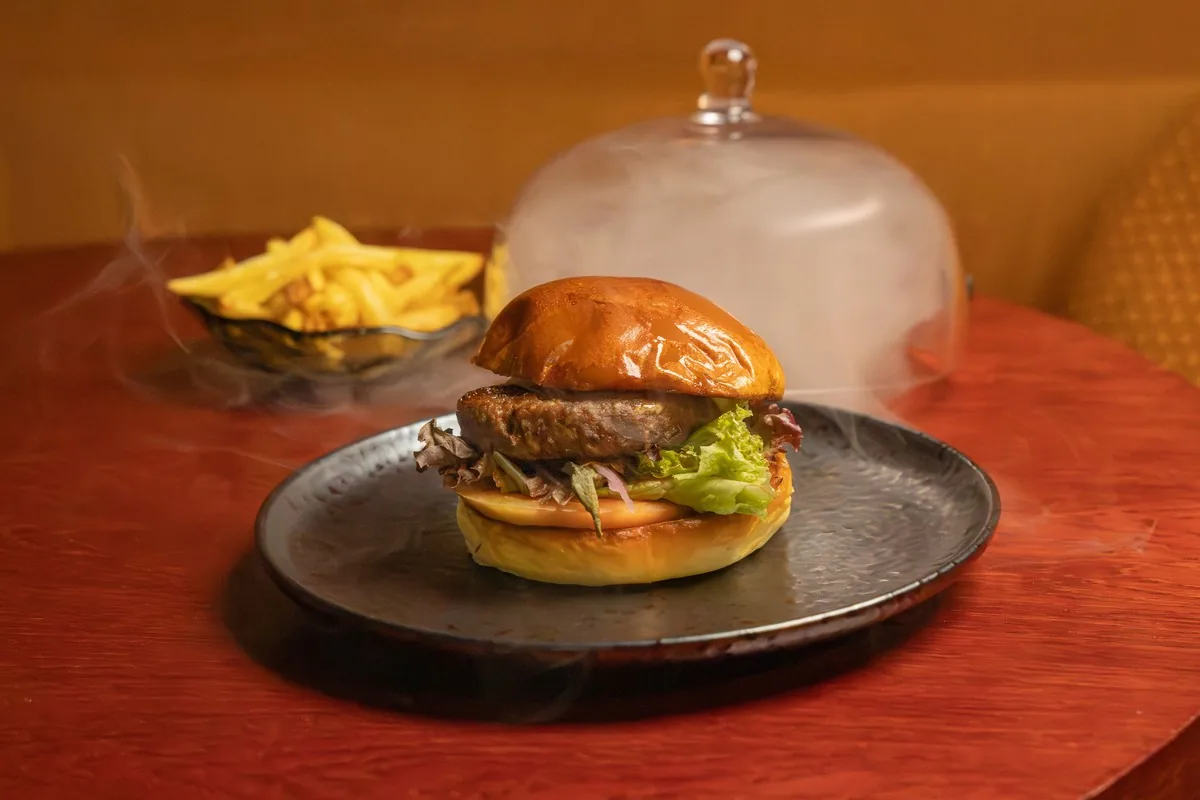 A burger with lettuce and tomato on a plate with a side of fries, served under a glass cloche