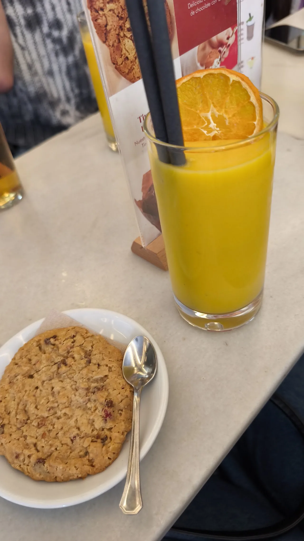 A glass of orange juice with a straw beside a cookie on a saucer