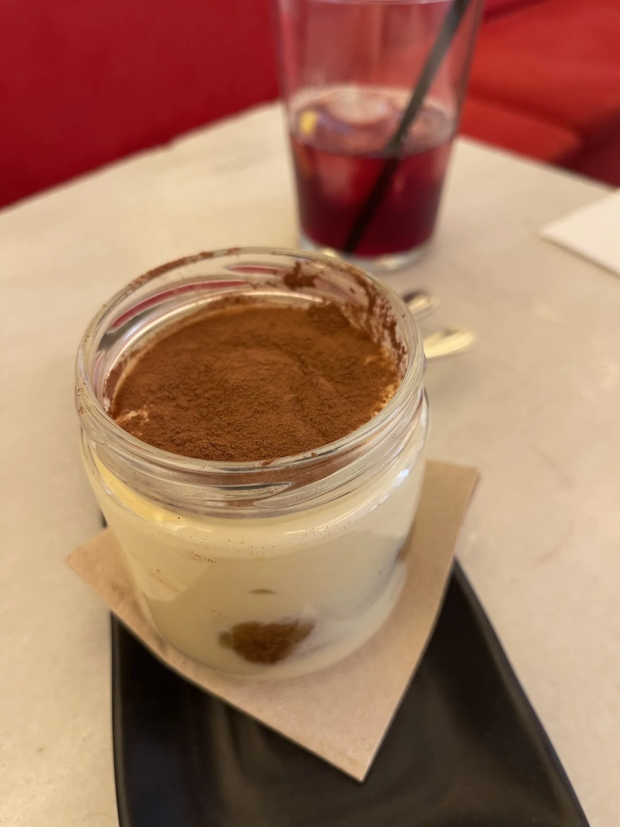 A tiramisu in a glass jar dusted with cocoa, beside a red drink