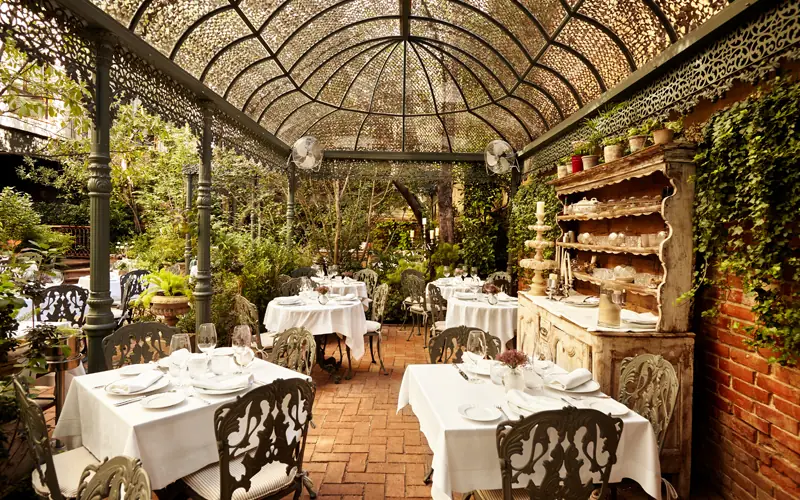 Covered garden terrace with lattice canopy, set tables in white linens, and an antique wooden sideboard