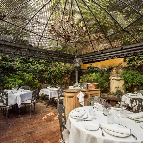Garden terrace under a lattice dome with a hanging chandelier, ivy-covered brick walls, and round set tables