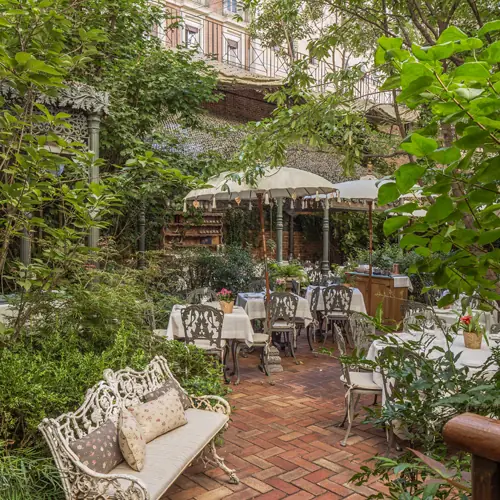 Leafy garden courtyard with umbrellas, a bench, and set tables surrounded by potted plants