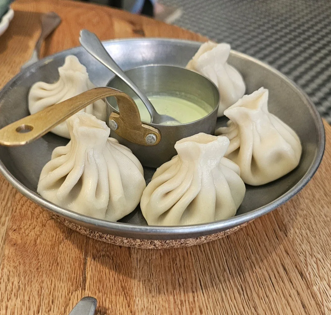 Six pleated dumplings in a metal dish with a small bowl of sauce