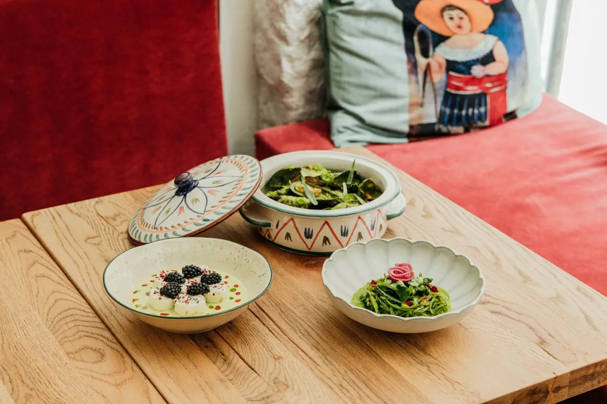 Three dishes on a table: cheese balls with blackberries, greens in a covered ceramic pot, and a shaved vegetable salad