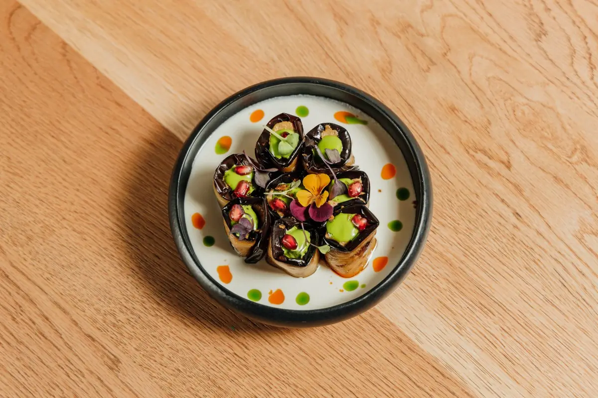 Rolled eggplant pieces filled with green paste, topped with pomegranate seeds and an edible flower on a black-rimmed plate