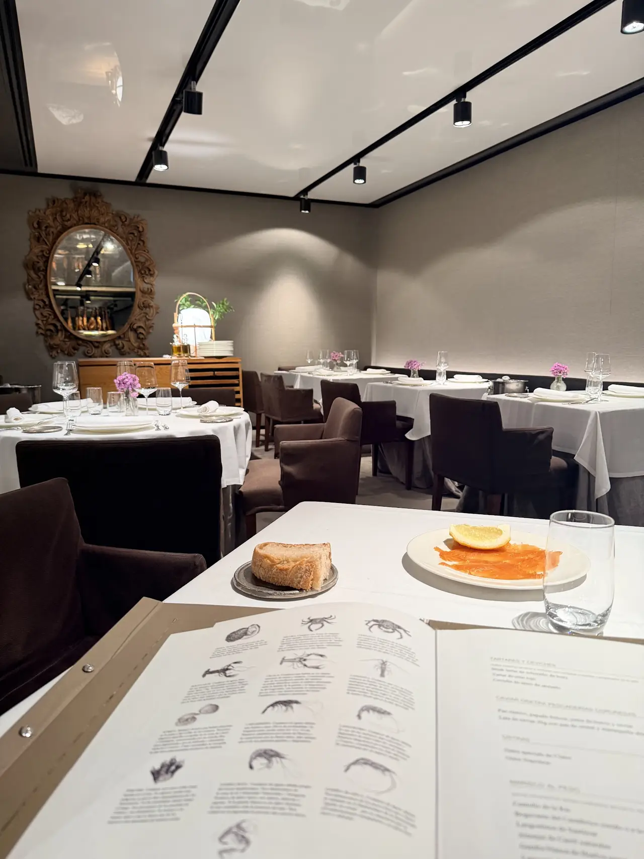 Table with bread, a plate of cured orange-colored fish and lemon, open menu, dining room behind