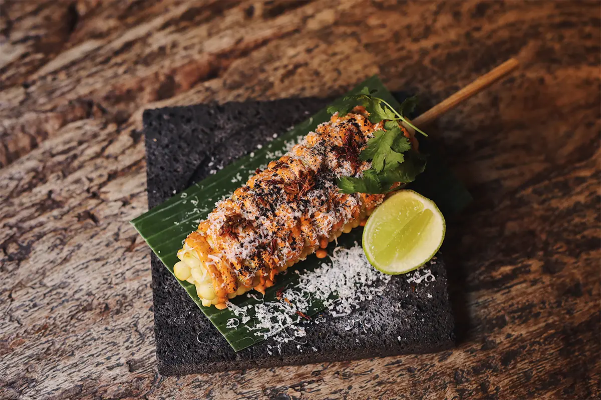 Grilled corn on the cob topped with crema, cotija cheese, chile powder, and cilantro, served on a banana leaf with a lime wedge, at Oaxaca
