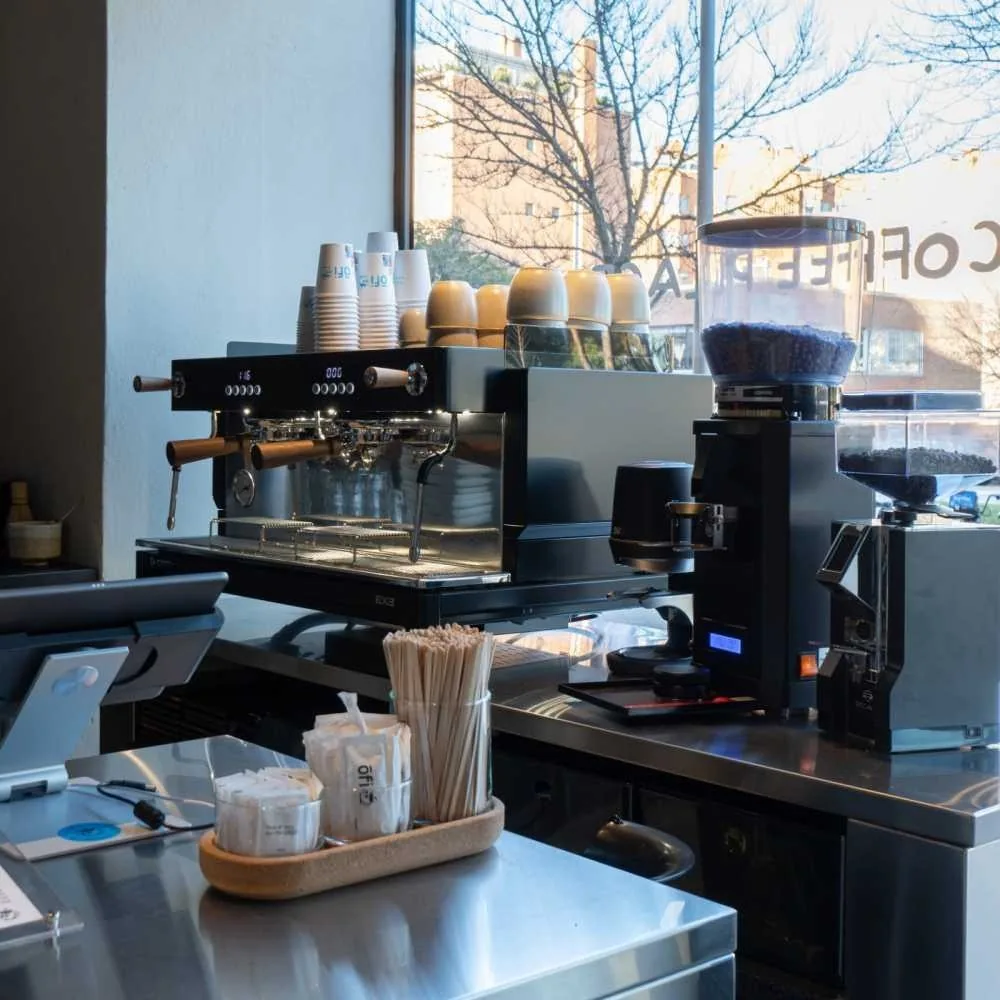 An espresso machine and grinder on a stainless steel cafe counter with stacked paper cups