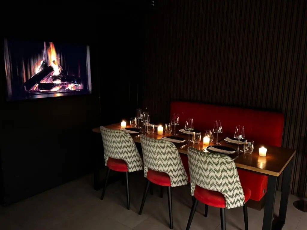 Intimate dining nook with a red banquette, patterned chairs with red seats, a candlelit set table and a wall screen showing a fireplace