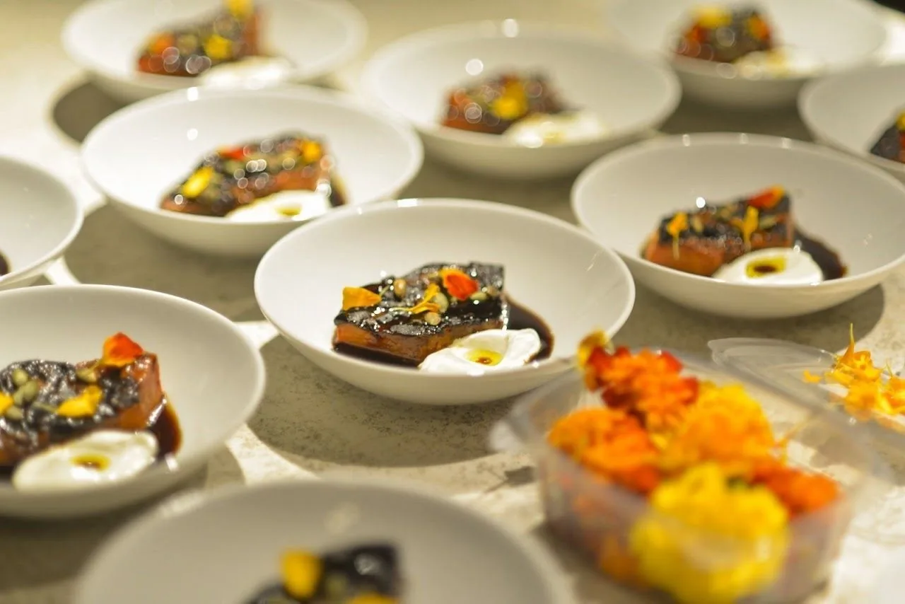 Rows of white bowls each holding a glazed piece of meat with cream and edible flowers