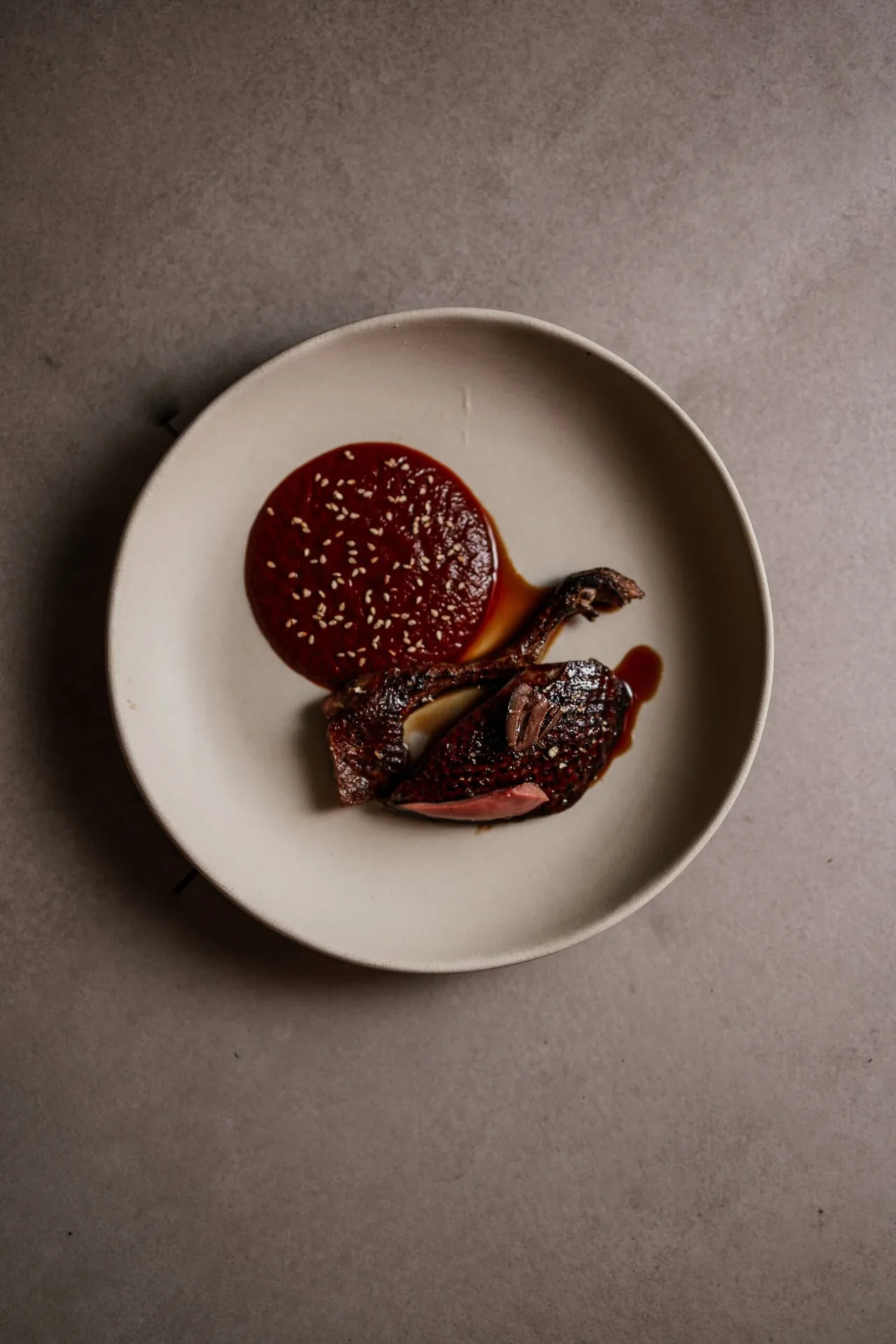 A round beige plate with a dark red sauce and a piece of meat on a grey surface