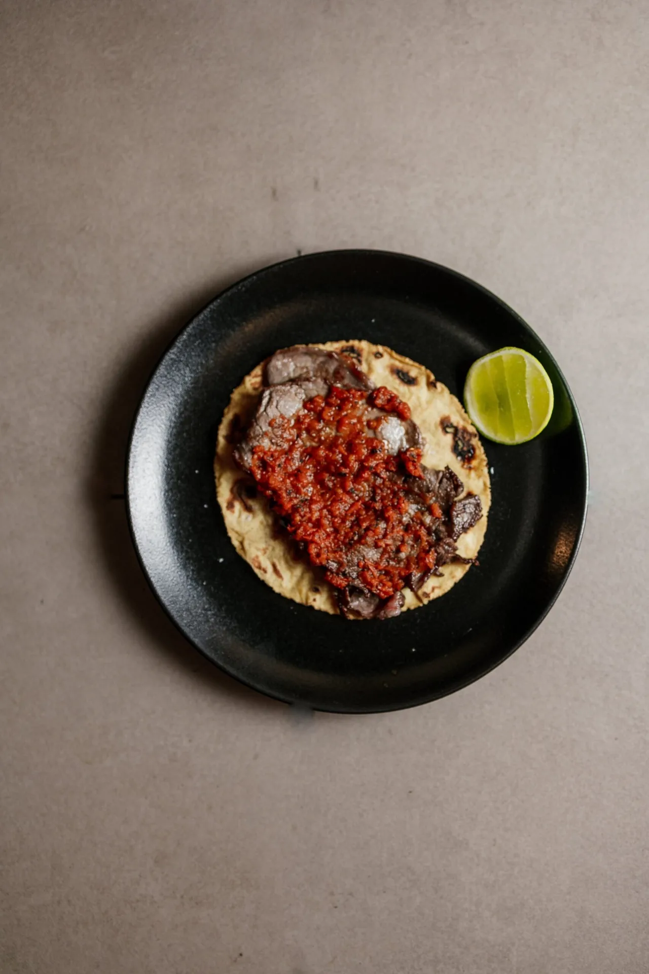 A round tostada topped with red sauce and meat on a black plate with a lime wedge