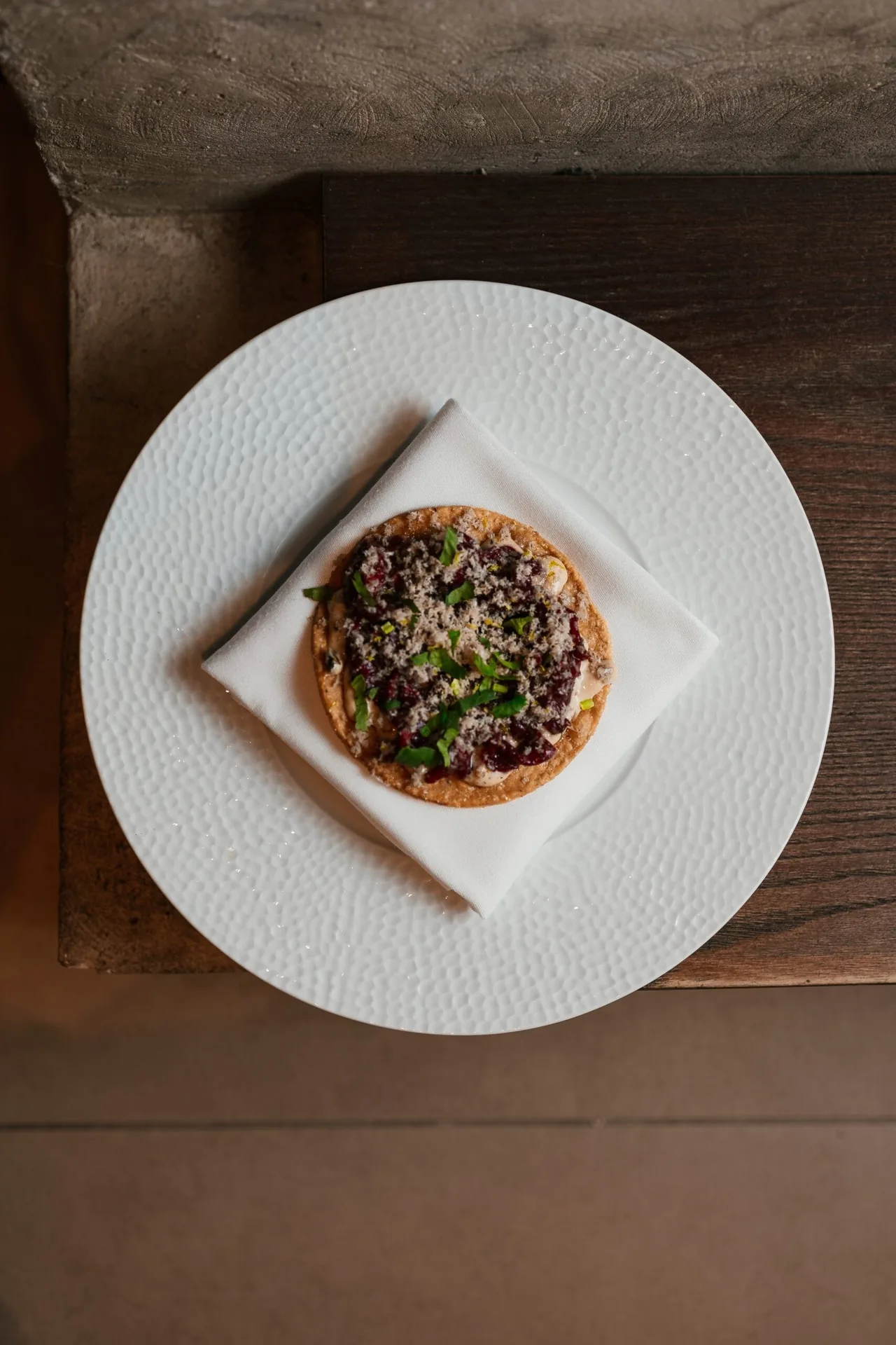 A small round tostada topped with dark meat on a white plate on a folded napkin