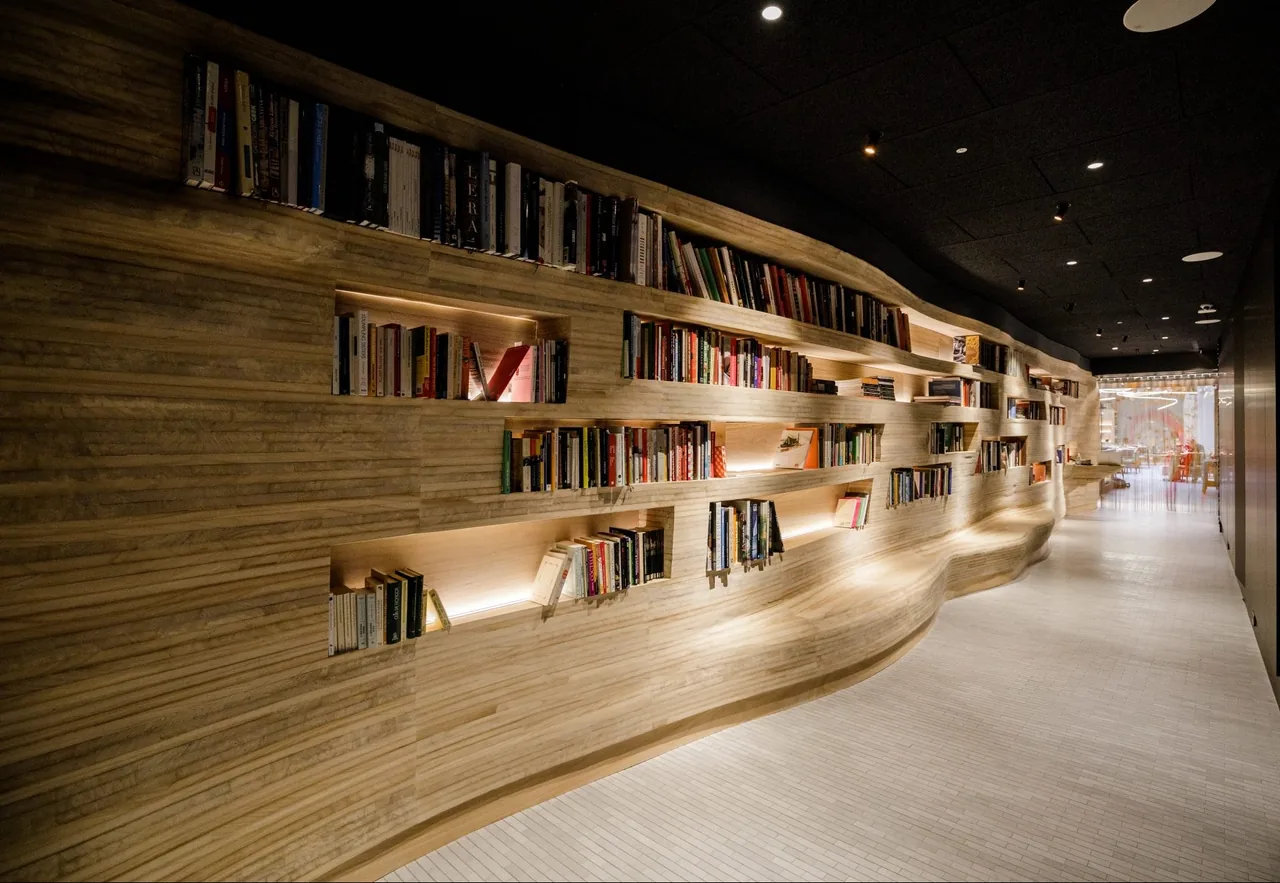 Curving sculpted pale-timber wall lined with lit recessed shelves of cookbooks, forming a corridor that leads toward the dining room