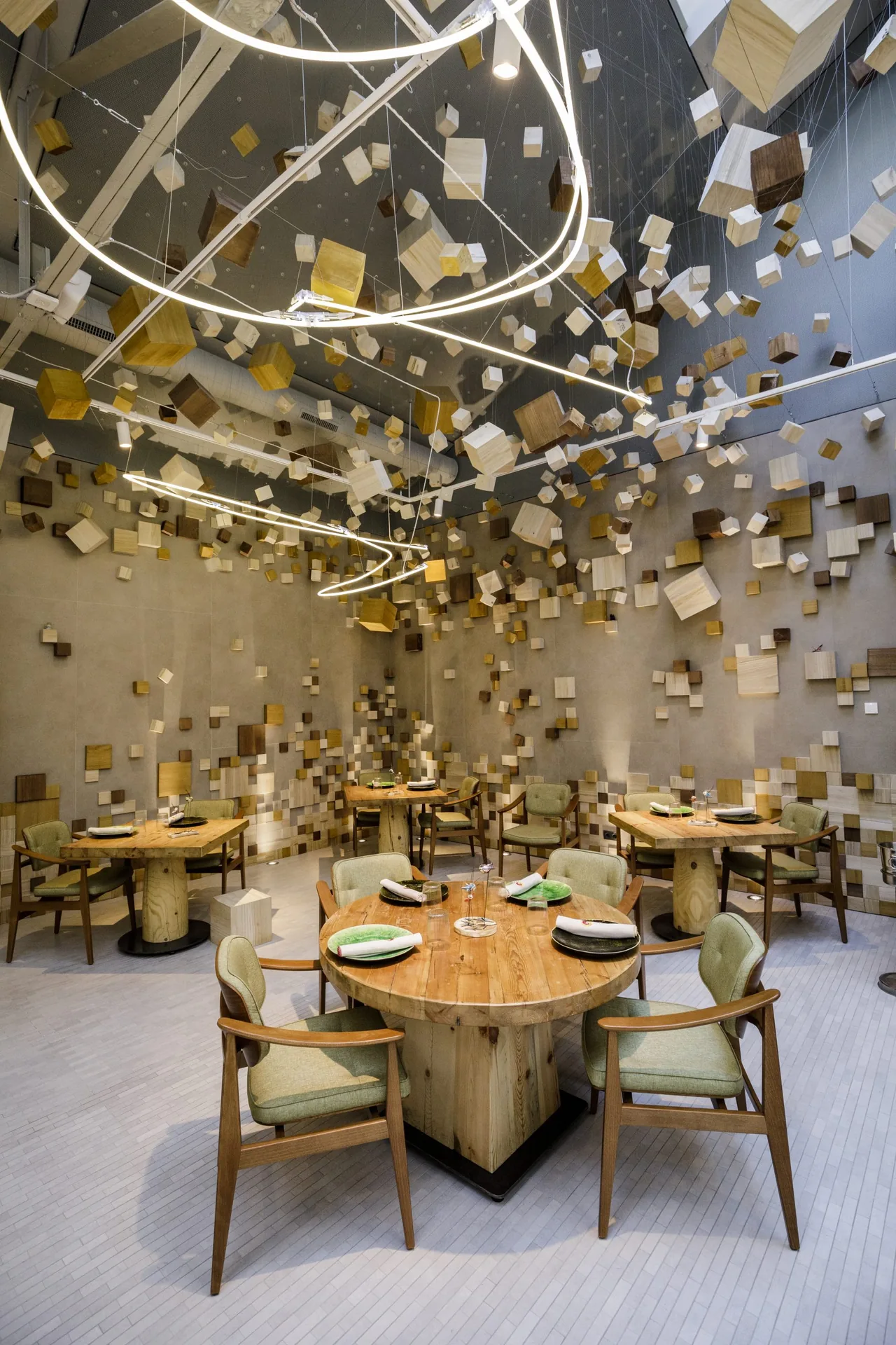 High-ceilinged dining room where hundreds of small wooden cubes hang on wires from the ceiling and stud the walls, above round timber tables set with green plates and sage-green armchairs