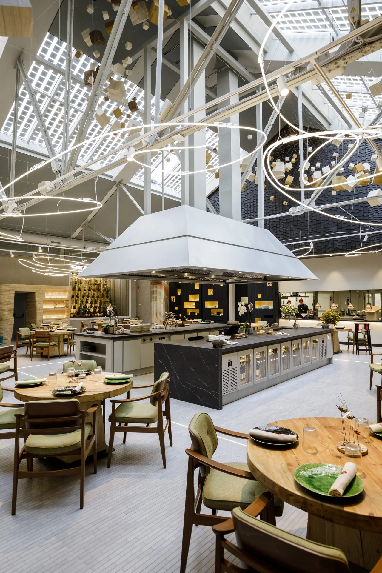 Skylit dining hall with a central open kitchen under a large white extractor canopy, cooks working at the pass beyond, and round wooden tables set with green plates in the foreground