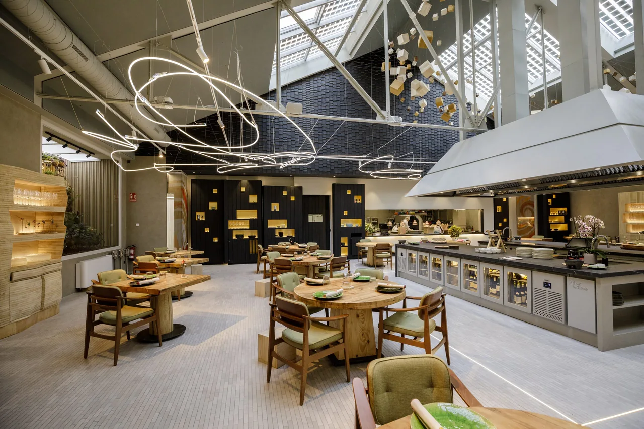 Wide view of the dining hall with looping neon-tube light sculptures overhead, sage-green armchairs at round timber tables, and the dark stone service counter with a chef at the pass on the right