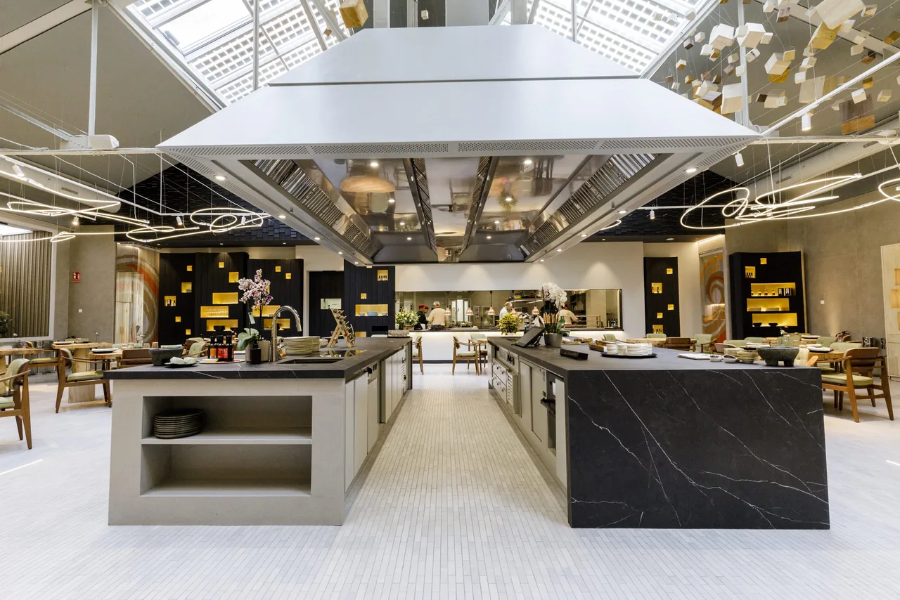 Head-on view of the twin dark-stone service counters under a suspended steel canopy, stacked plates and orchids on top, with cooks at the kitchen pass in the background