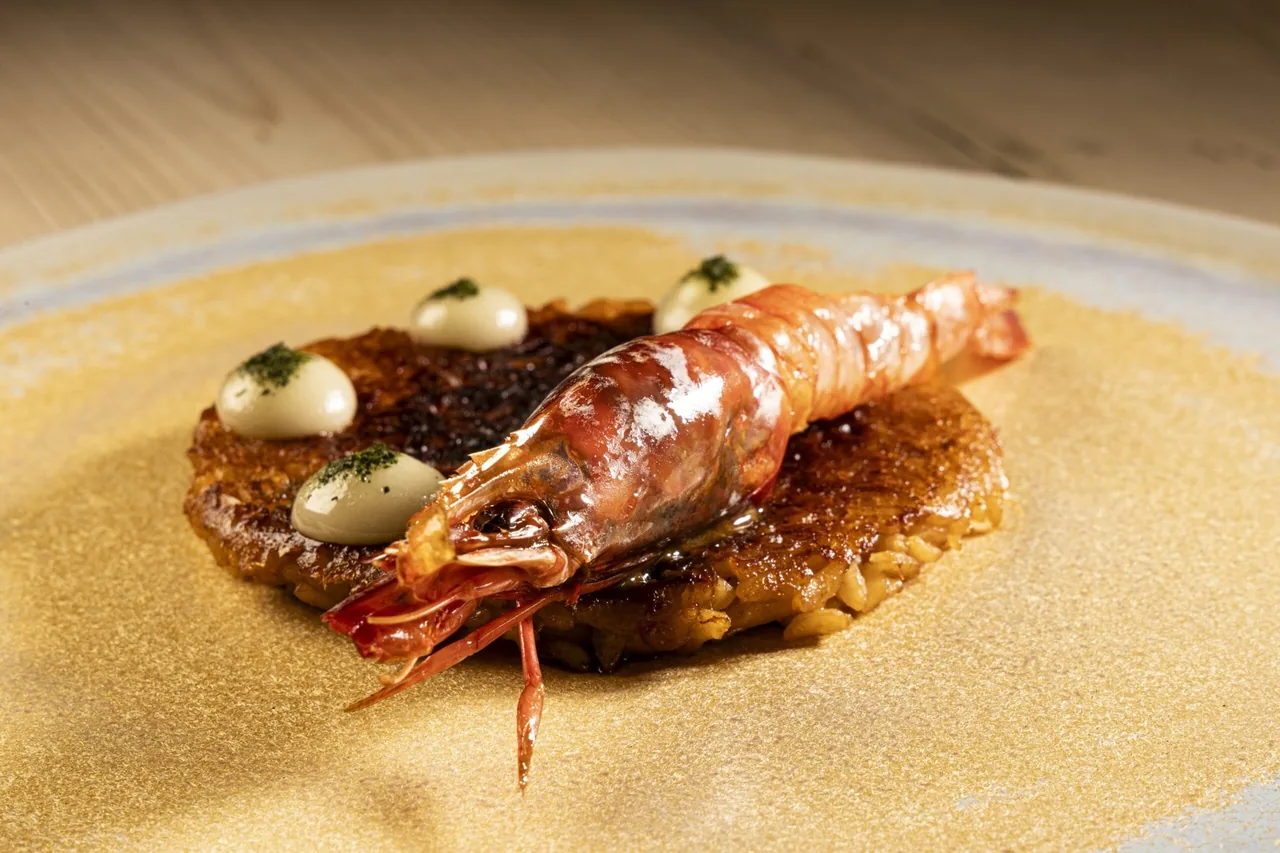 A red prawn resting on a crisp rice cake with dots of white cream on a golden plate