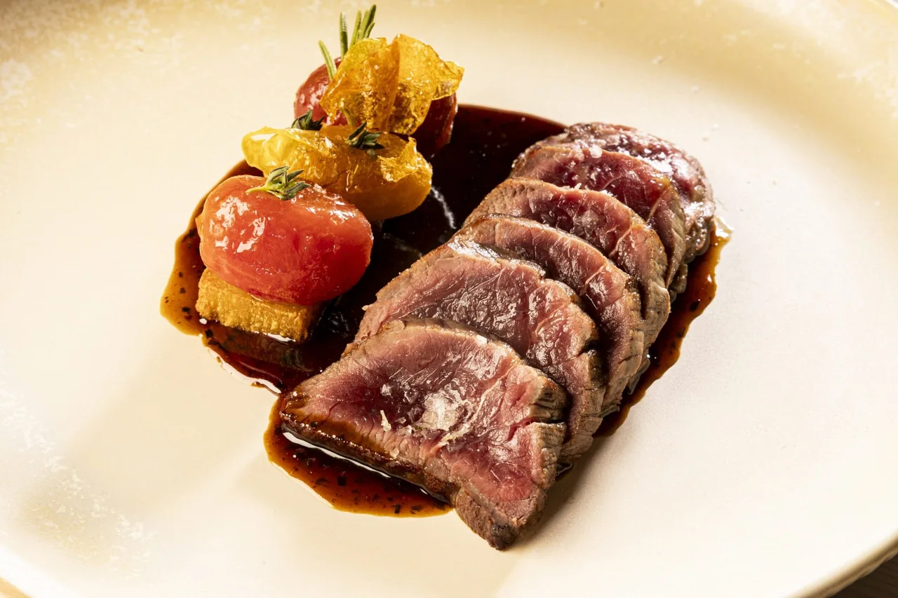 Slices of pink-centred meat on a cream plate with a glazed tomato, a crisp and a dark sauce