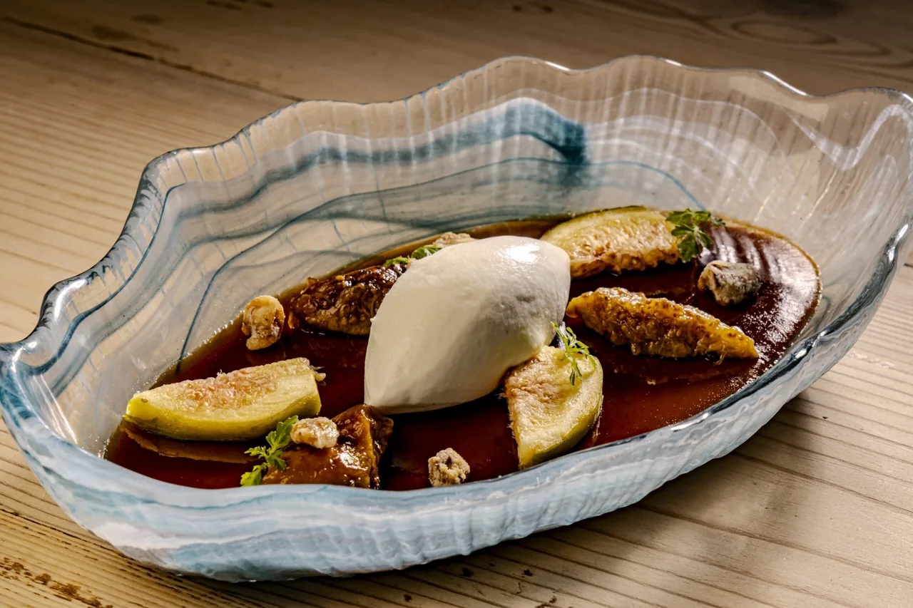 A glass bowl with a scoop of white cream, fig pieces and a dark sauce on a wooden table