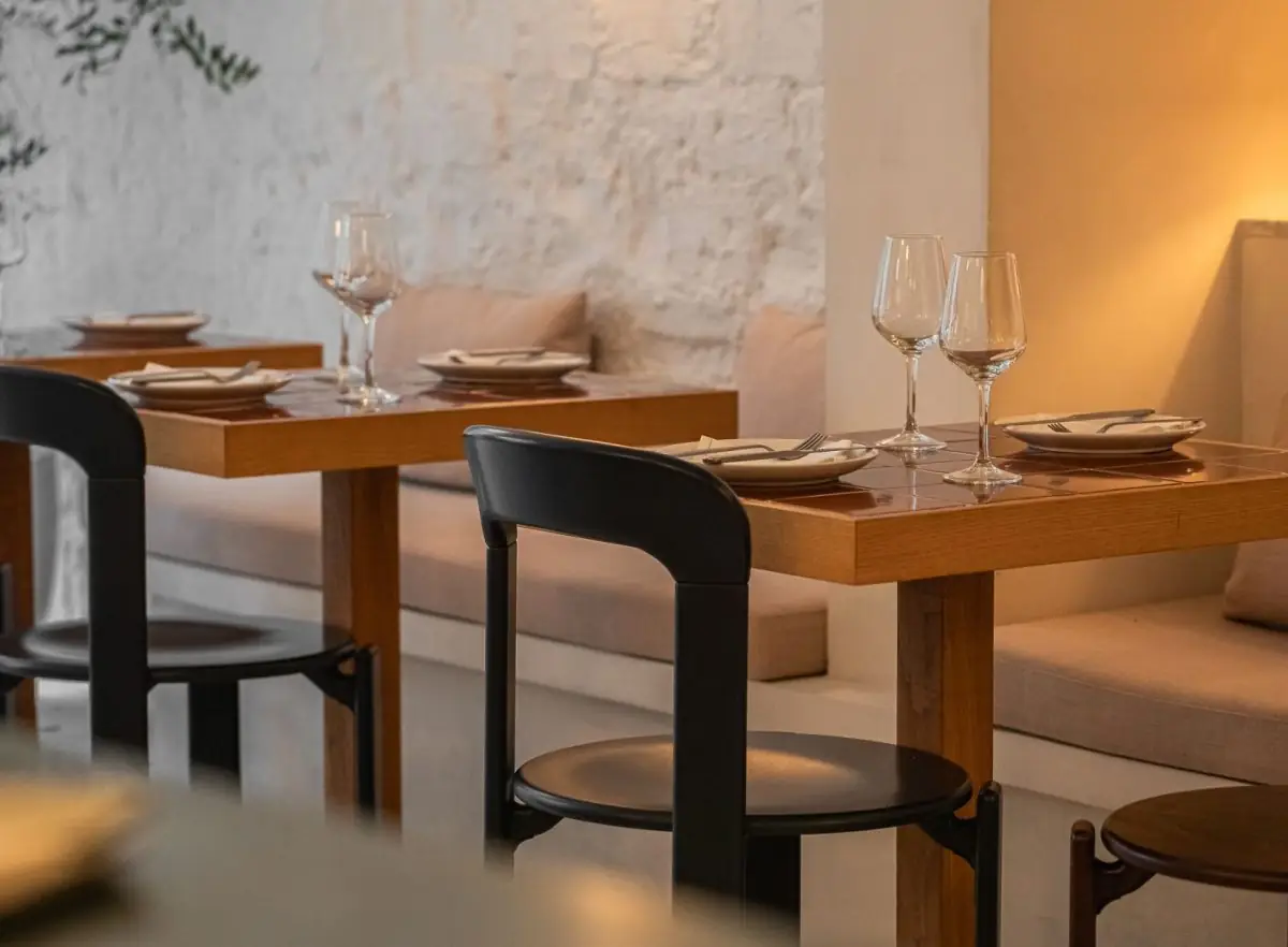 Warm-lit dining room with wooden tile-topped tables, black tubular chairs, upholstered banquettes, wine glasses and place settings against a whitewashed stone wall