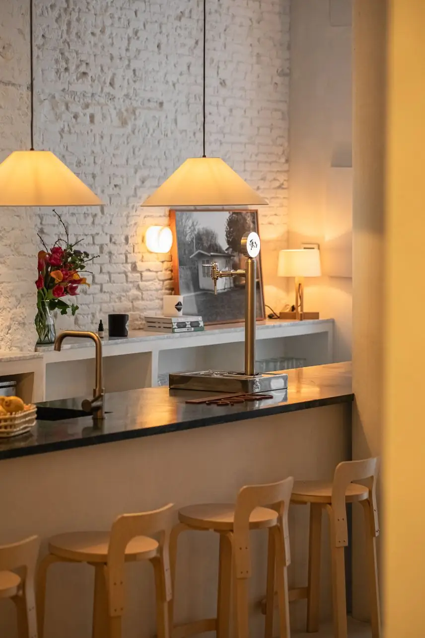 Bar counter with dark stone top, brass beer tap, blond wood stools and conical pendant lamps against an exposed whitewashed brick wall with fresh flowers