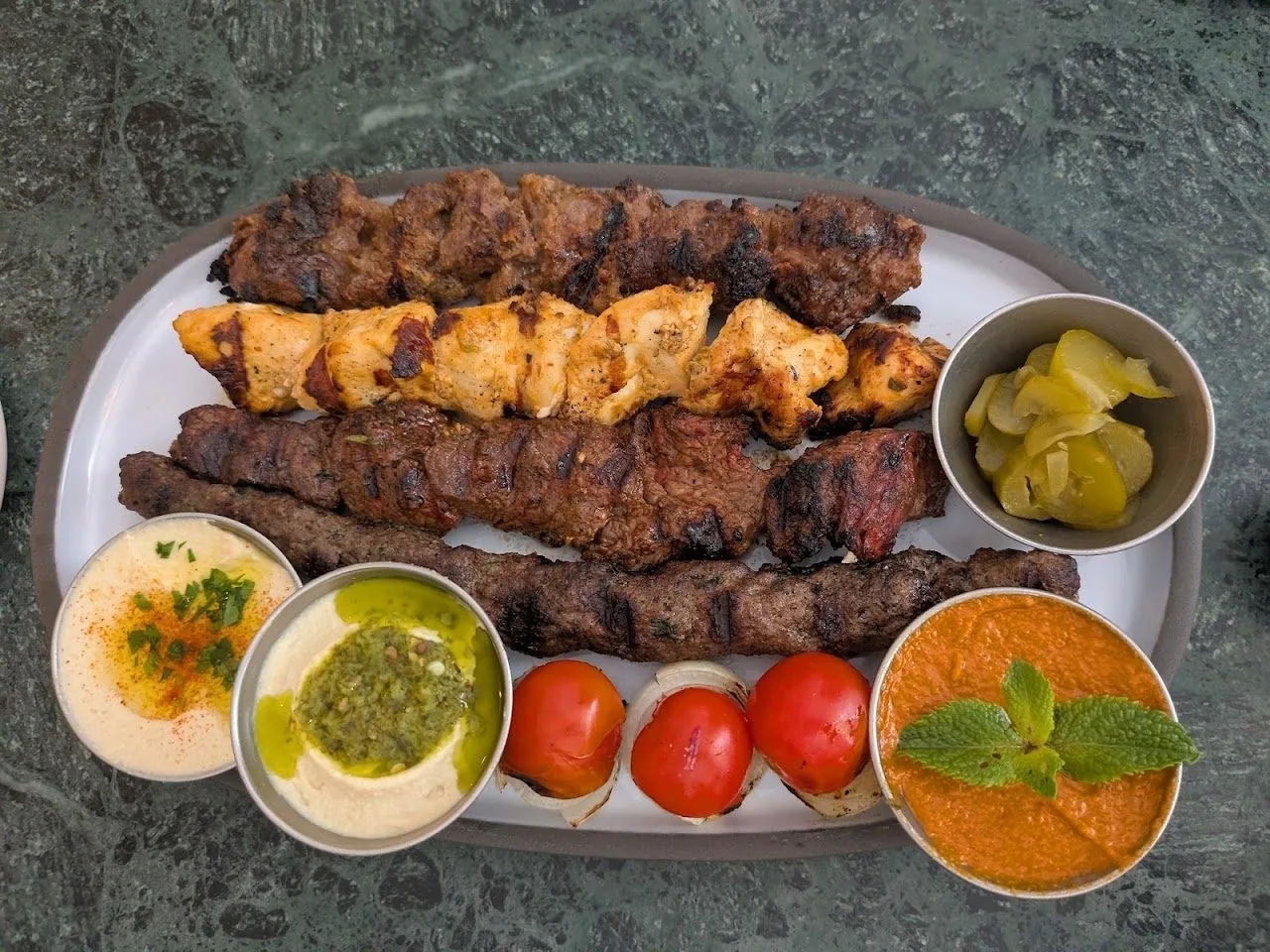 A grey platter of grilled kebabs with dips, pickles and tomatoes