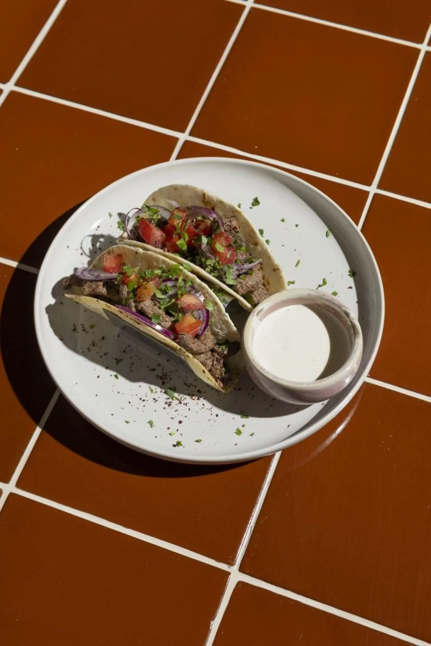 A plate of two filled tacos with a small pot of white sauce on a tiled surface