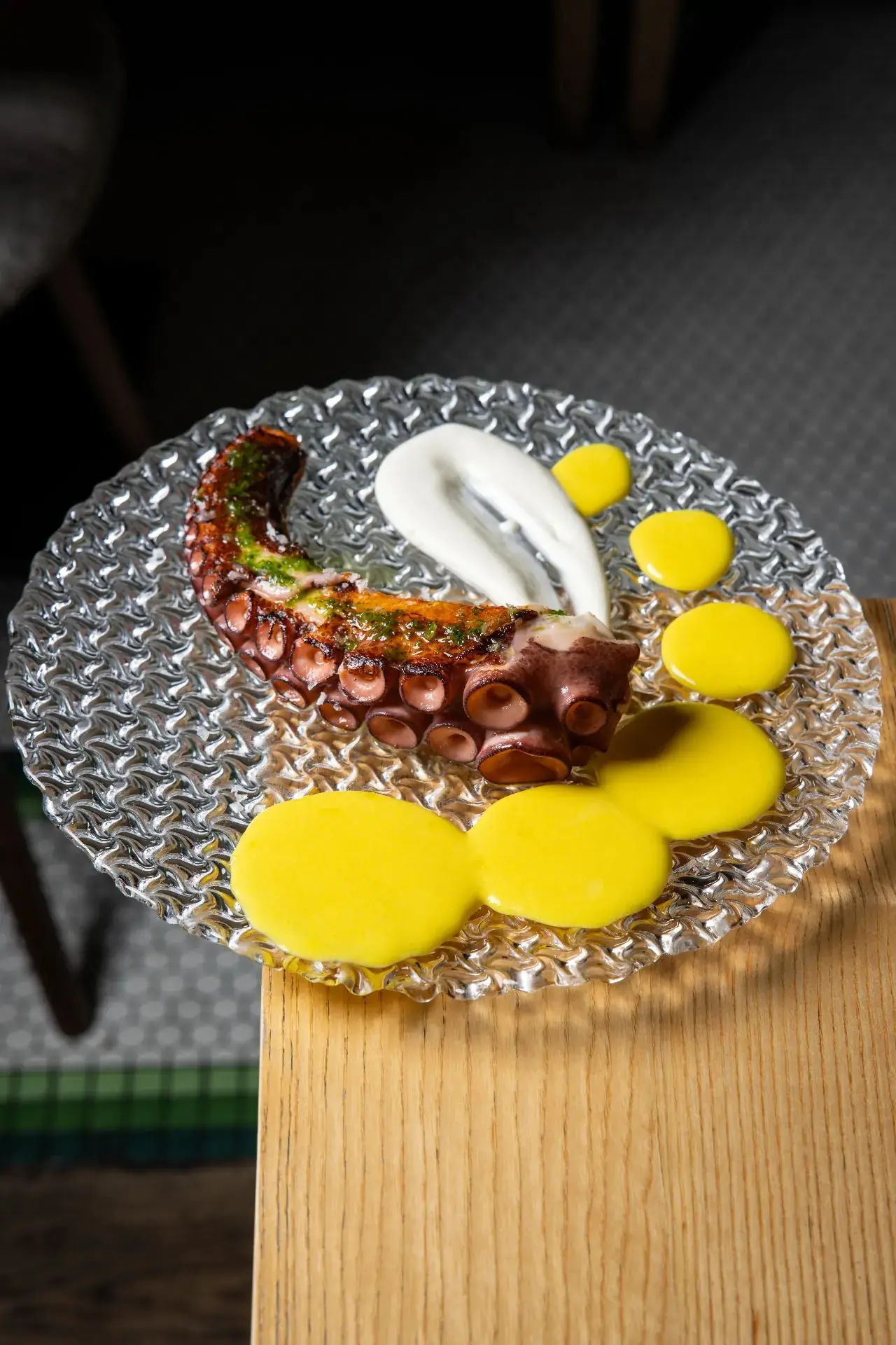 Charred grilled octopus tentacle with herb oil and a quenelle of foam, plated with saffron-yellow potato-cream dots on a textured clear glass plate