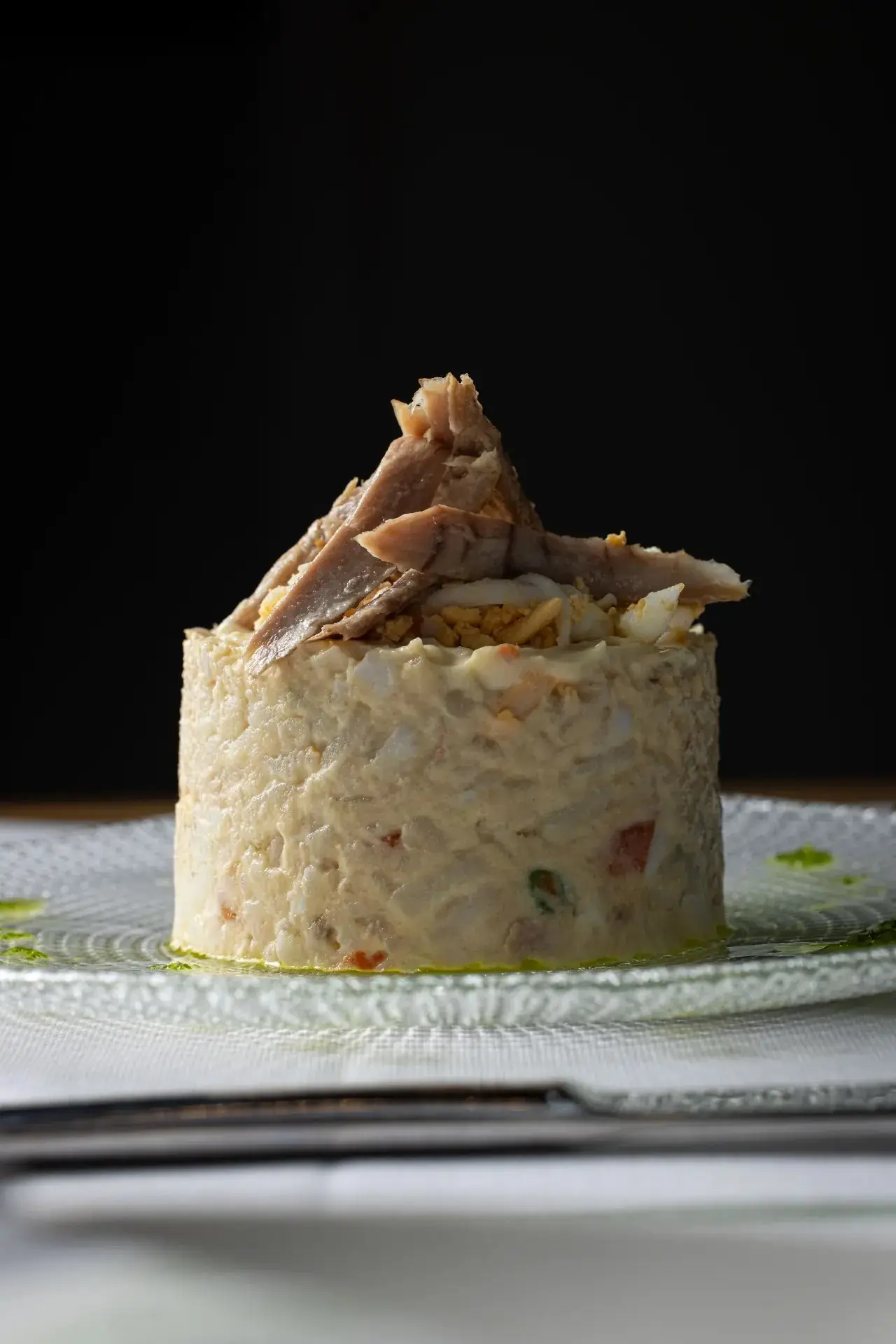 Cylindrical mound of ensaladilla rusa topped with flaked tuna belly and chopped egg on a textured glass plate against a dark background
