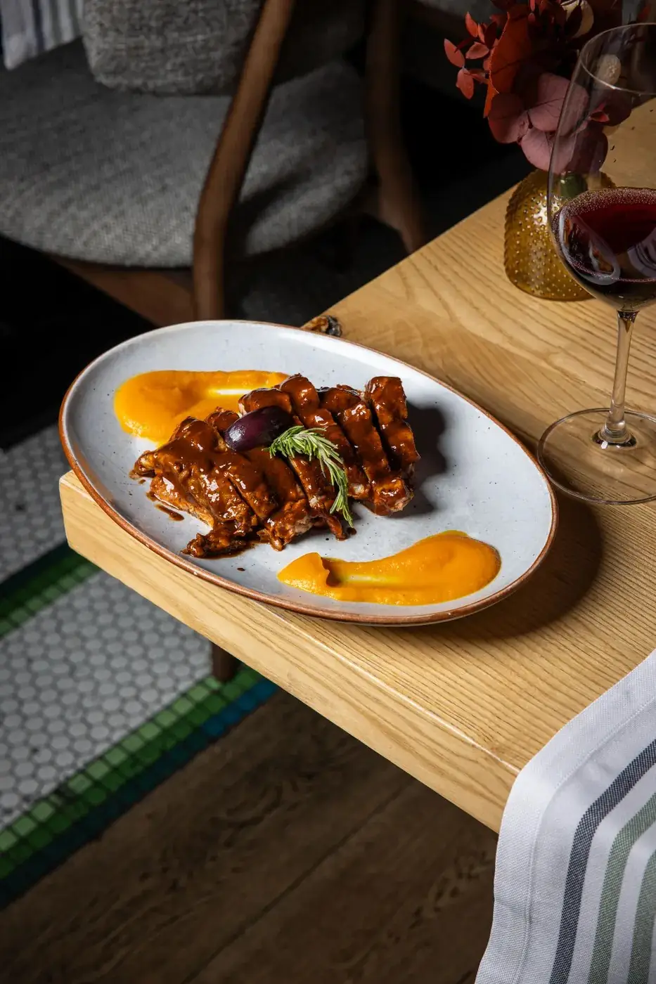 Sliced glazed meat in dark sauce with two smears of orange squash puree and a rosemary sprig on an oval speckled plate, a glass of red wine alongside on a wooden table