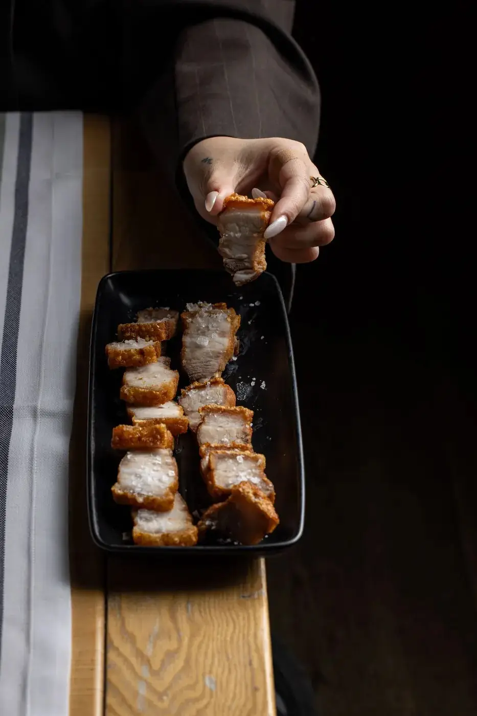 Cubes of crisp-skinned fried pork belly dusted with flaked salt in a black rectangular dish, a hand lifting one piece