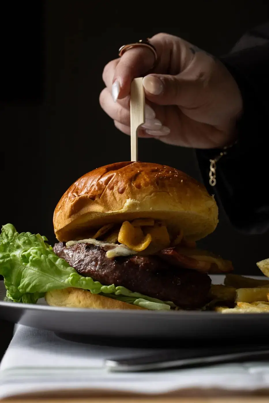 Burger with a thick beef patty, lettuce and brioche bun served with fries on a grey plate, a hand inserting a wooden skewer