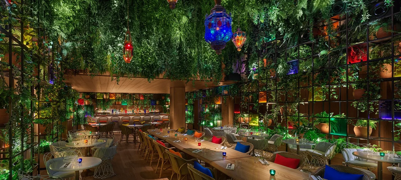 Dining room walled in dense green foliage with coloured backlit niches, hanging Moroccan lanterns, a long communal table with bright cushions and a lit bar at the back