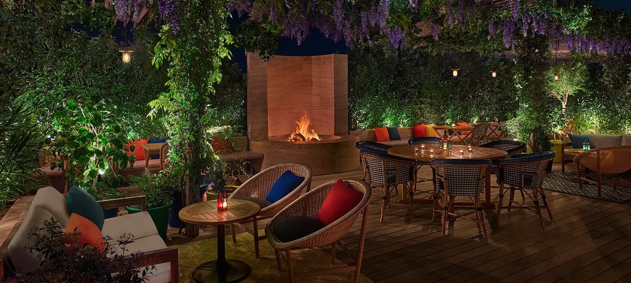 The same planted terrace at night, a fire burning in a tall stone hearth, round tables lit by lanterns and candles under wisteria