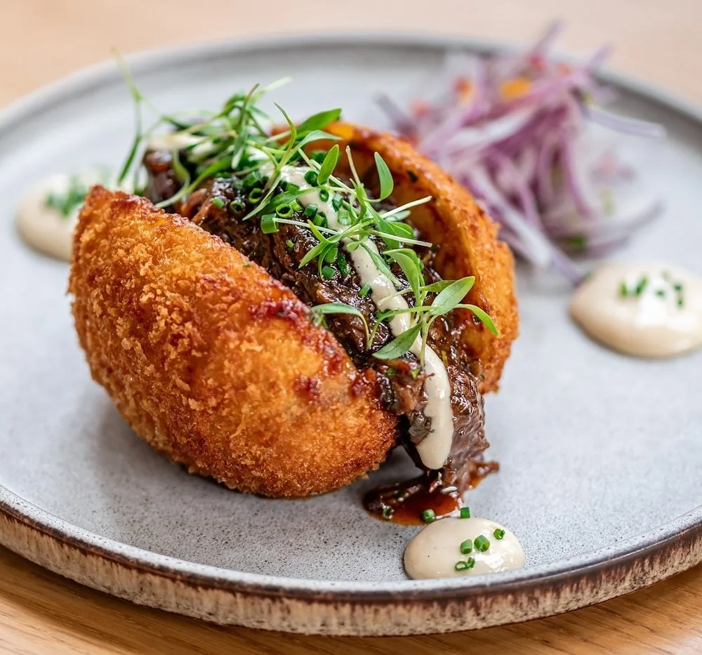 A golden breaded croquette split open and filled with braised meat and herbs, with red onion and sauce on a grey plate
