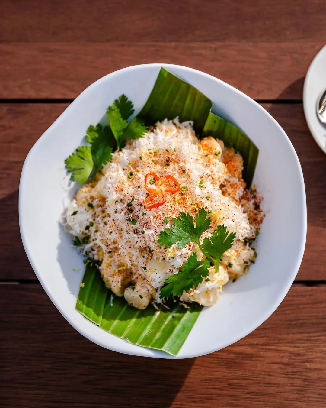 A white bowl of pale kernels topped with grated cheese, chilli and coriander on a banana leaf