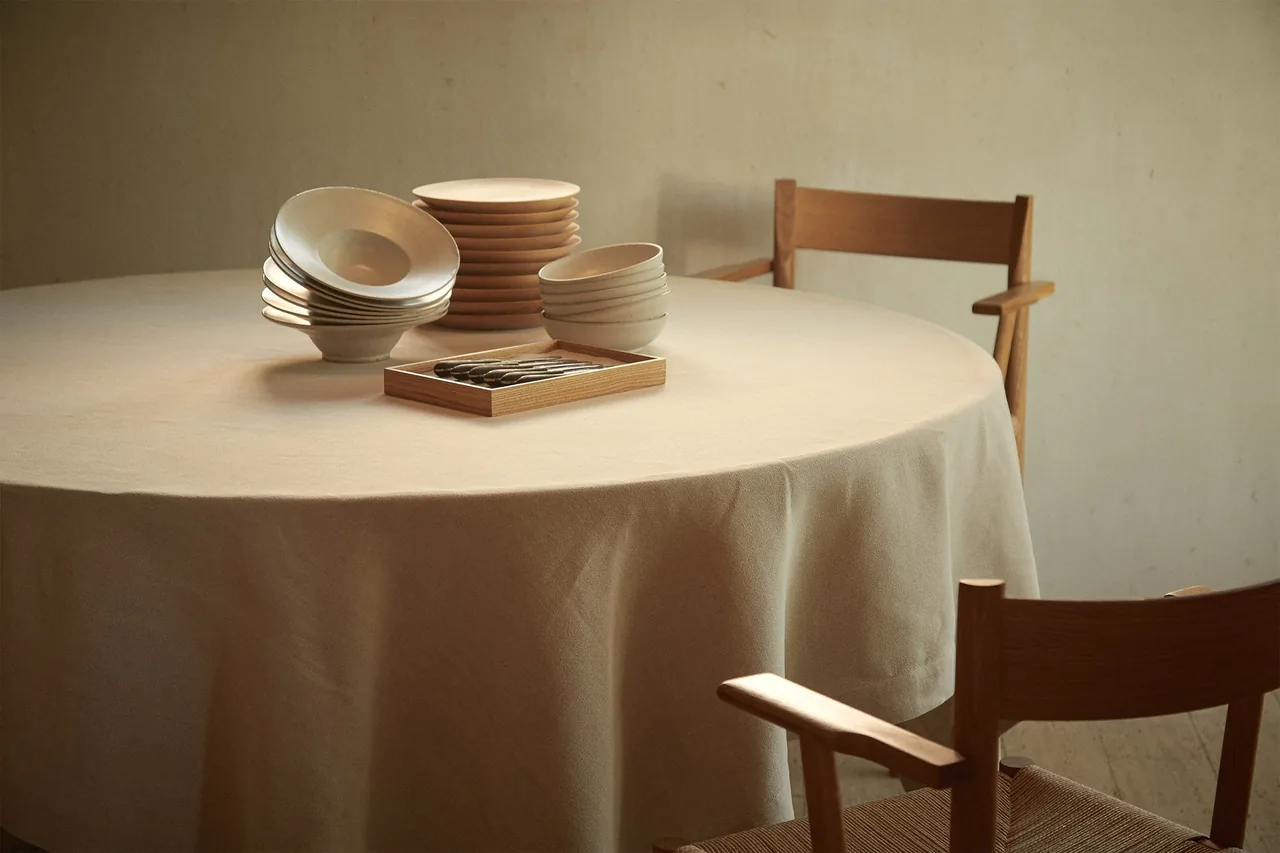 A round table with a linen cloth, a stack of ceramic bowls and wooden chairs against a grey wall