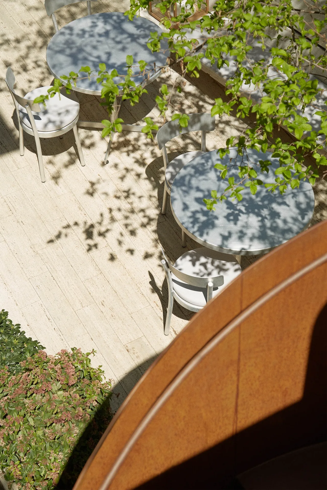 A round table and chairs on a pale stone terrace seen from above, with leaf shadows and a curved rust-coloured wall